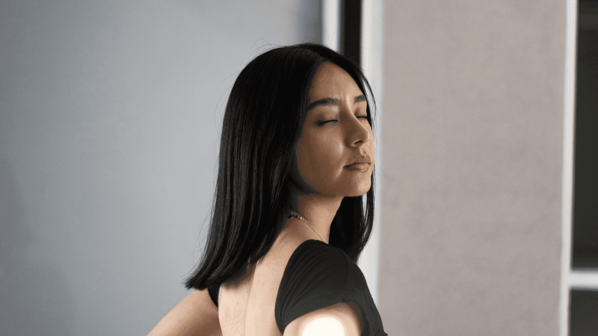 Woman with long dark hair in a black top, eyes closed, against a neutral background.